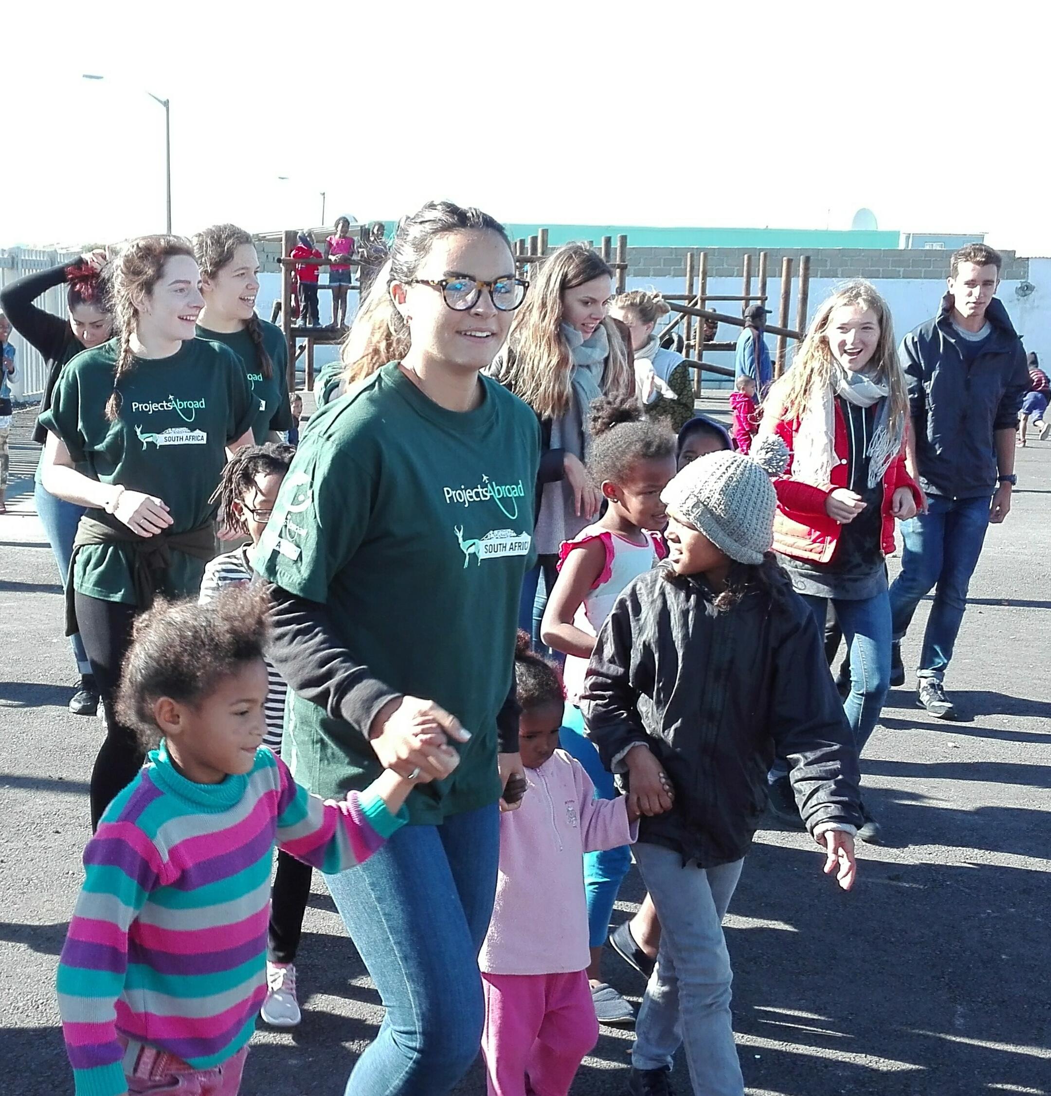 Projects Abroad volunteers take local children on a walk through the village at a Childcare Project in South Africa. Female volunters with children in South Africa hold hands of local children and lead them on a walk at their Childcare Project.